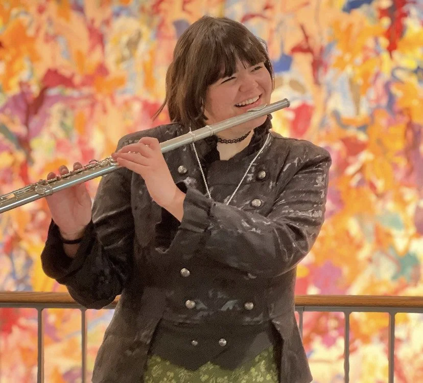 A young woman with short dark hair playing a silver flute. She is smiling and wearing a black jacket with metallic buttons, a black choker, and a necklace. The background is vibrant with colorful autumnal foliage.