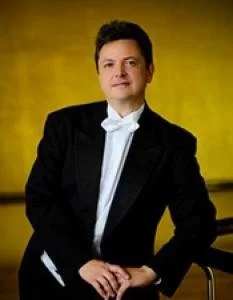 Portrait of a man in a tuxedo with a white bow tie, standing against a yellow background.