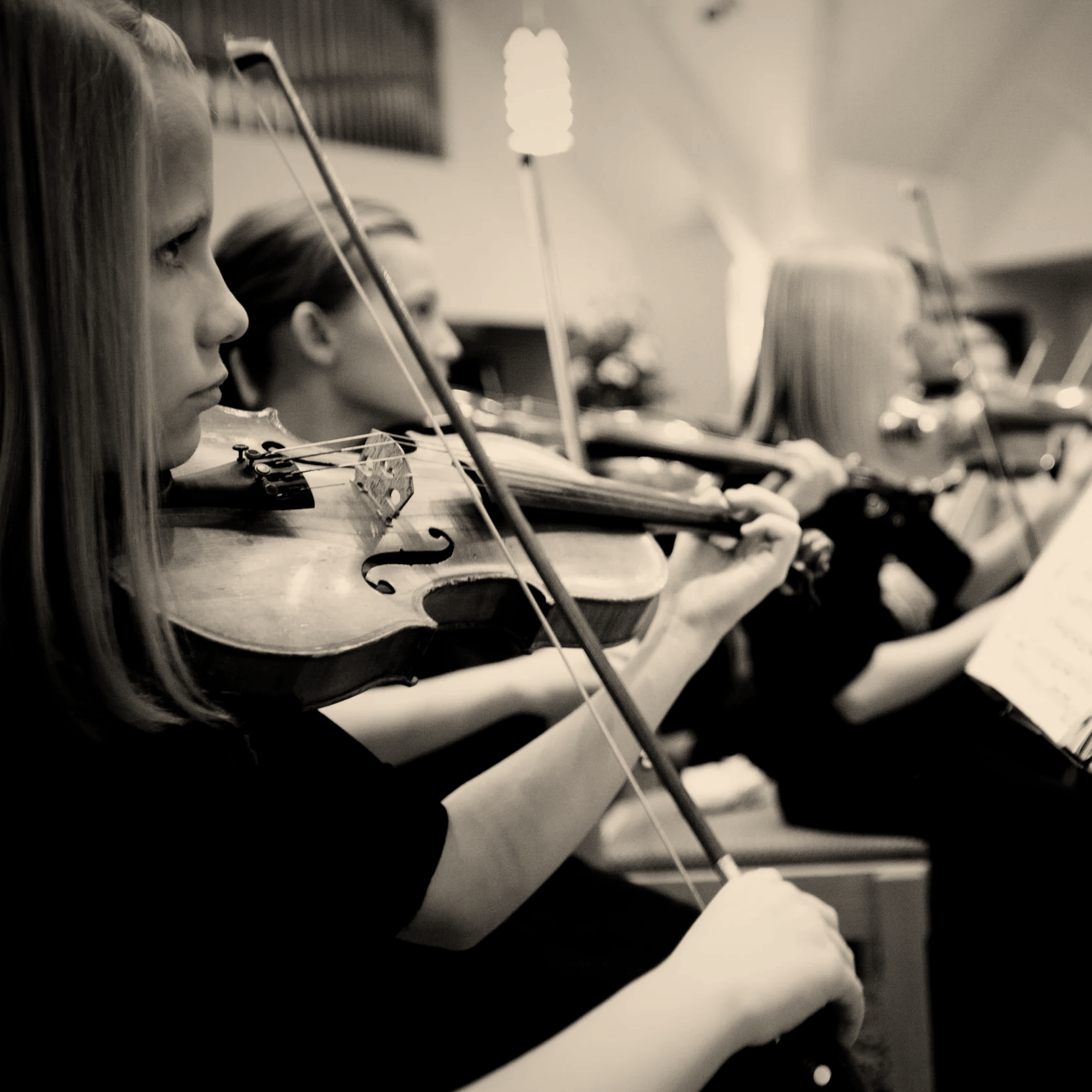 Women playing violins in an orchestra, focused on their instruments, in a black and white photo.