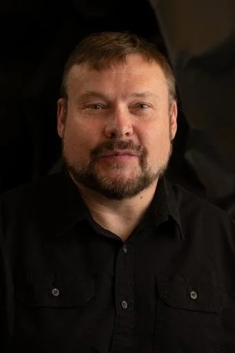 A man with short brown hair and a beard, wearing a black shirt, looking at the camera against a dark background.
