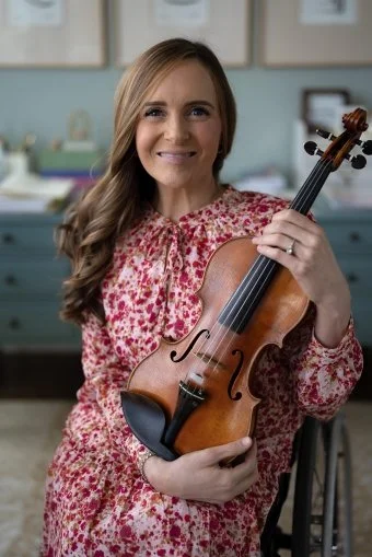 A woman smiling and holding a violin in a room with blue cabinets and picture frames on the wall.