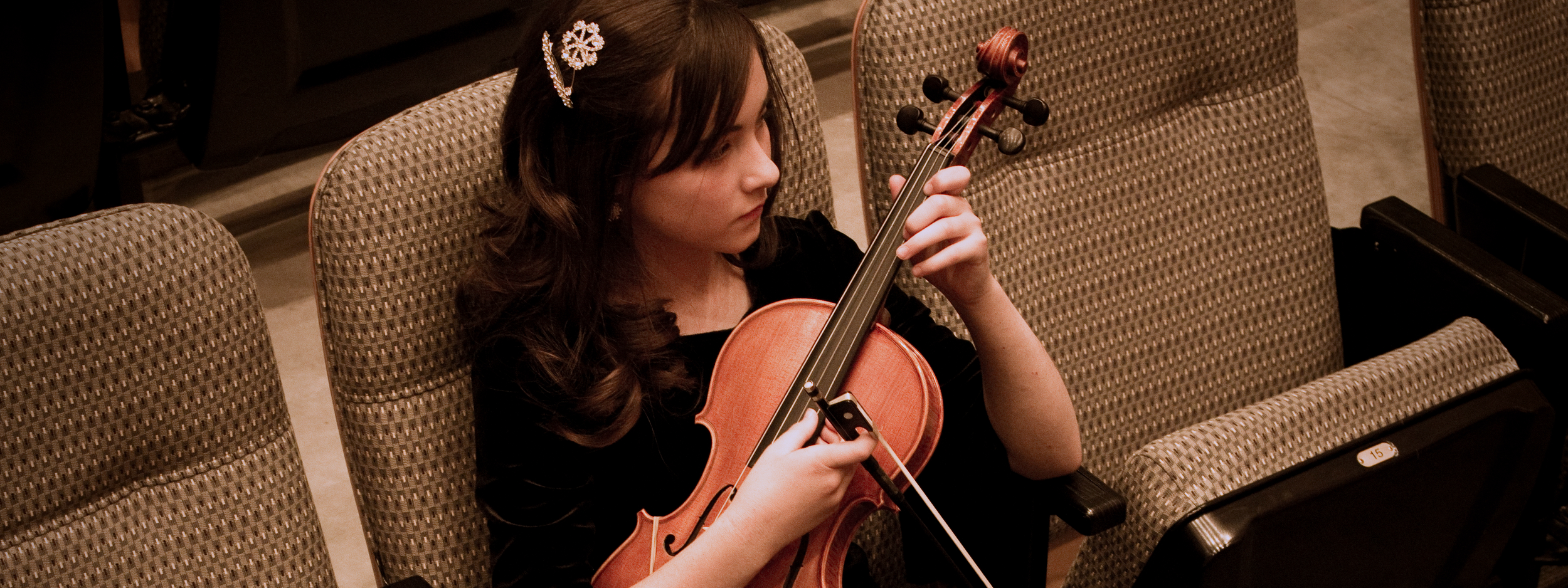 A young girl with dark hair adorned with a flower-themed hair clip, playing a violin while seated in a concert hall with beige patterned chairs.