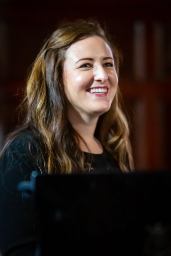 Smiling woman with long, wavy brown hair in a dimly lit indoor setting.