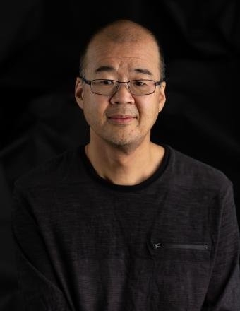 Portrait of a middle-aged man with glasses, wearing a black shirt, against a dark background.