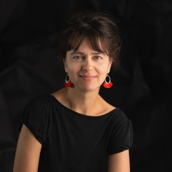 Woman with short dark hair wearing a black top and red earrings, smiling against a dark background.