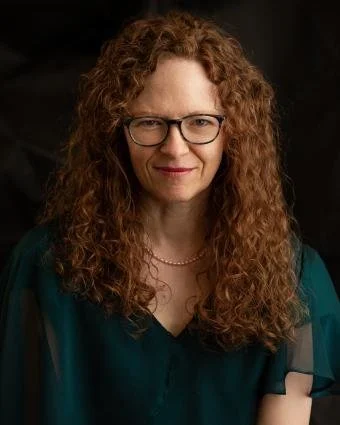 A woman with curly red hair, glasses, and a teal blouse, smiling in a professional portrait.