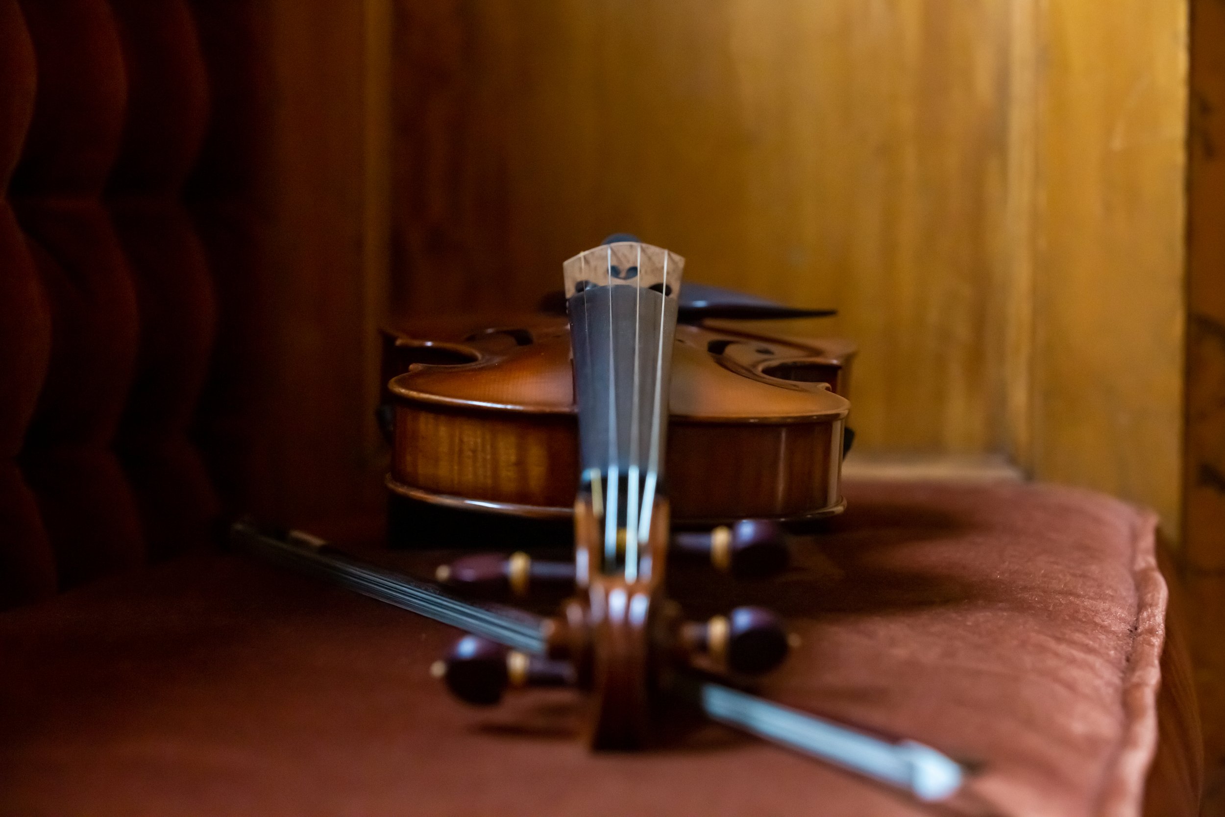 A close-up view of a violin bow resting on a wooden table, with several violins lined up in the background in a dimly lit room.