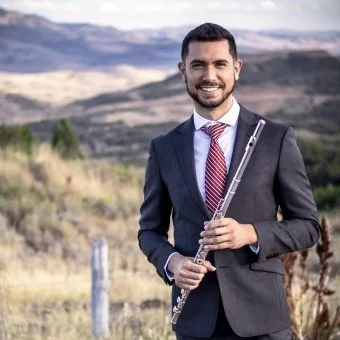 Man in a suit holding a flute outdoors with a landscape background.