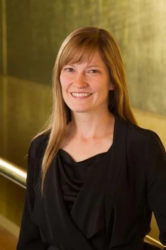 A smiling woman with long, light brown hair and bangs, wearing a black top, standing indoors with a yellow-green background.