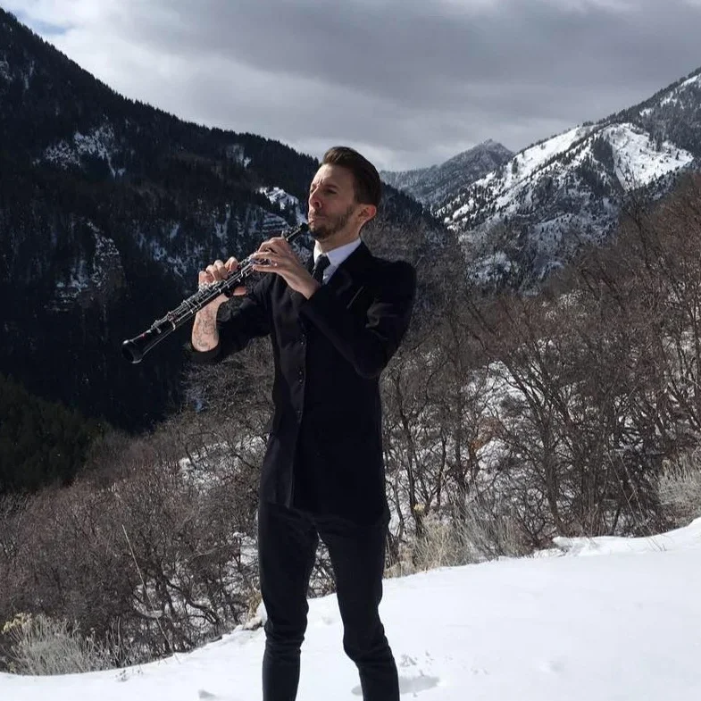 A man dressed in a black suit playing a clarinet outdoors in a snowy mountain landscape with cloudy sky.