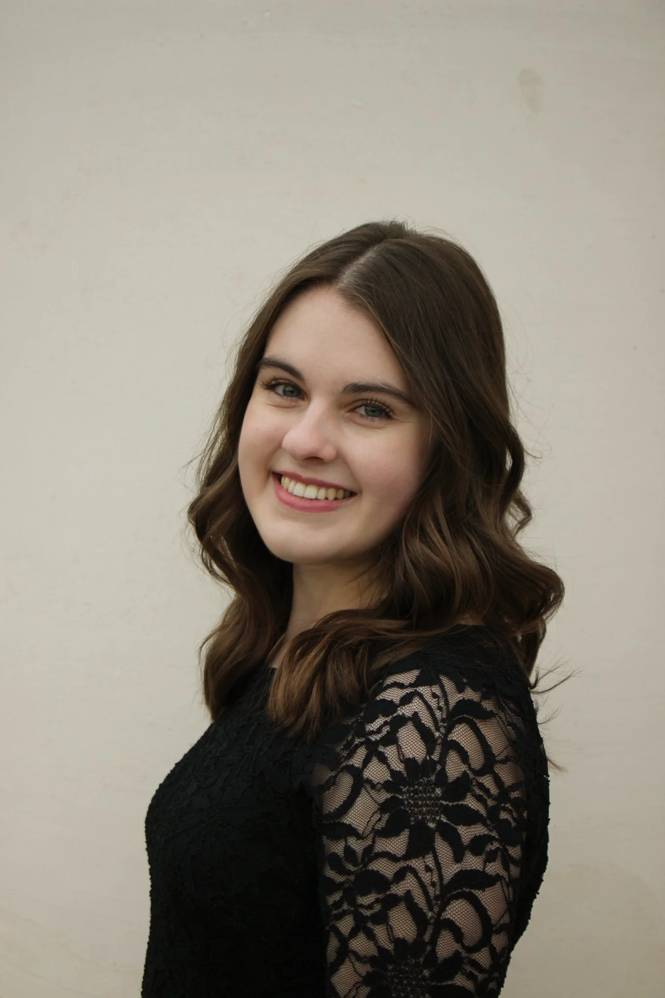 Young woman with shoulder-length wavy brown hair, smiling, wearing a black lace dress, standing against a plain beige wall.