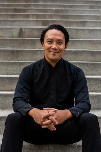 A man sitting on outdoor steps, smiling, wearing a black shirt with his hands clasped in front of him.
