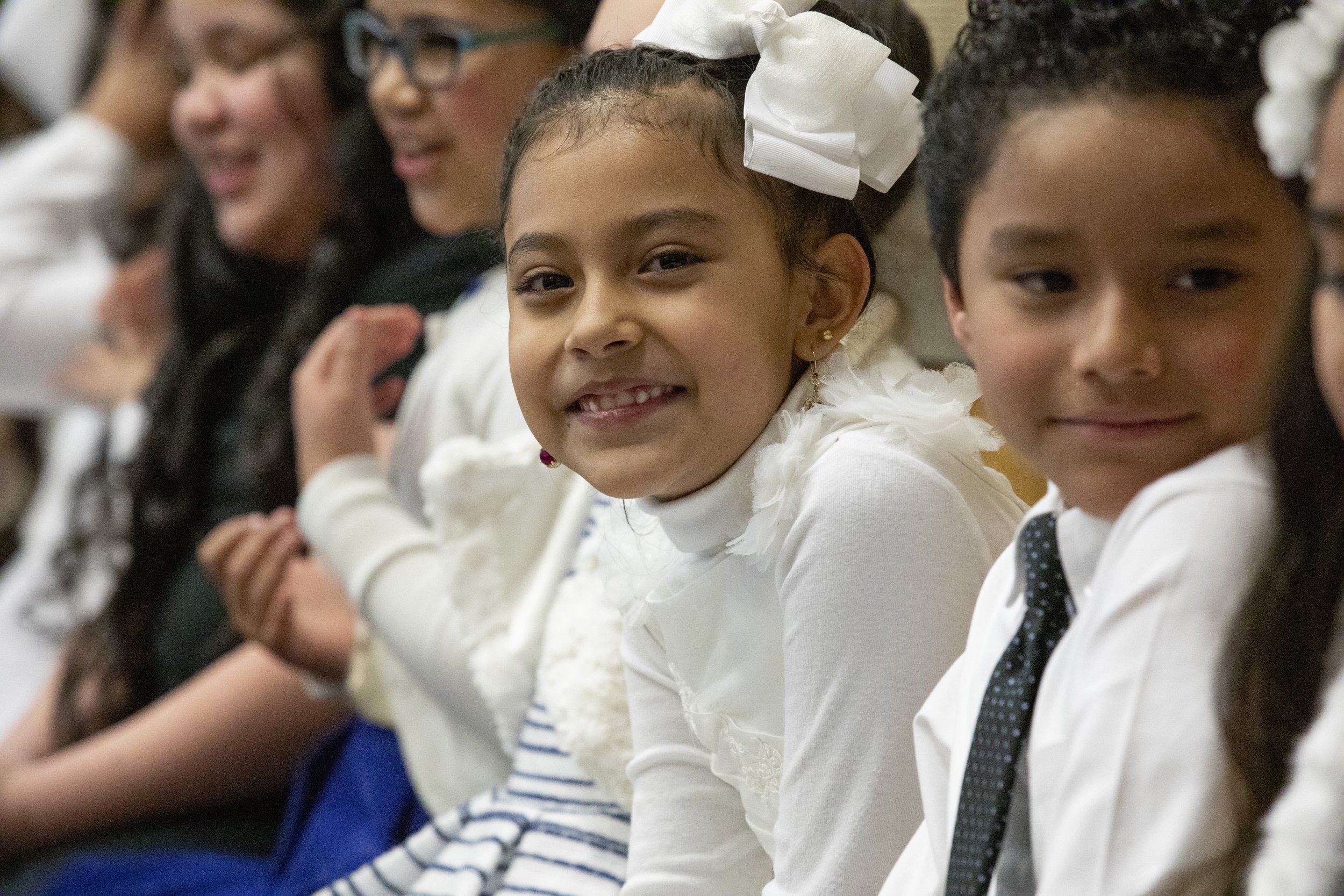Group of young children sitting in a row, dressed in formal attire, smiling and interacting at an event.