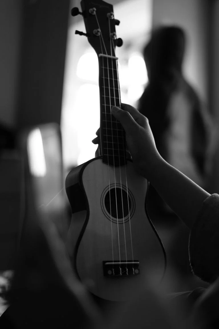 Person holding a small acoustic guitar with a hand on the fretboard, inside a room with natural light coming through windows, black and white photo.