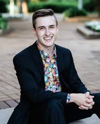 A young man in a floral shirt and blazer sitting outdoors on a brick pathway, smiling at the camera.
