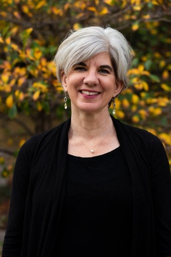 A woman with short gray hair smiling outdoors, standing in front of a tree with yellow and green leaves.