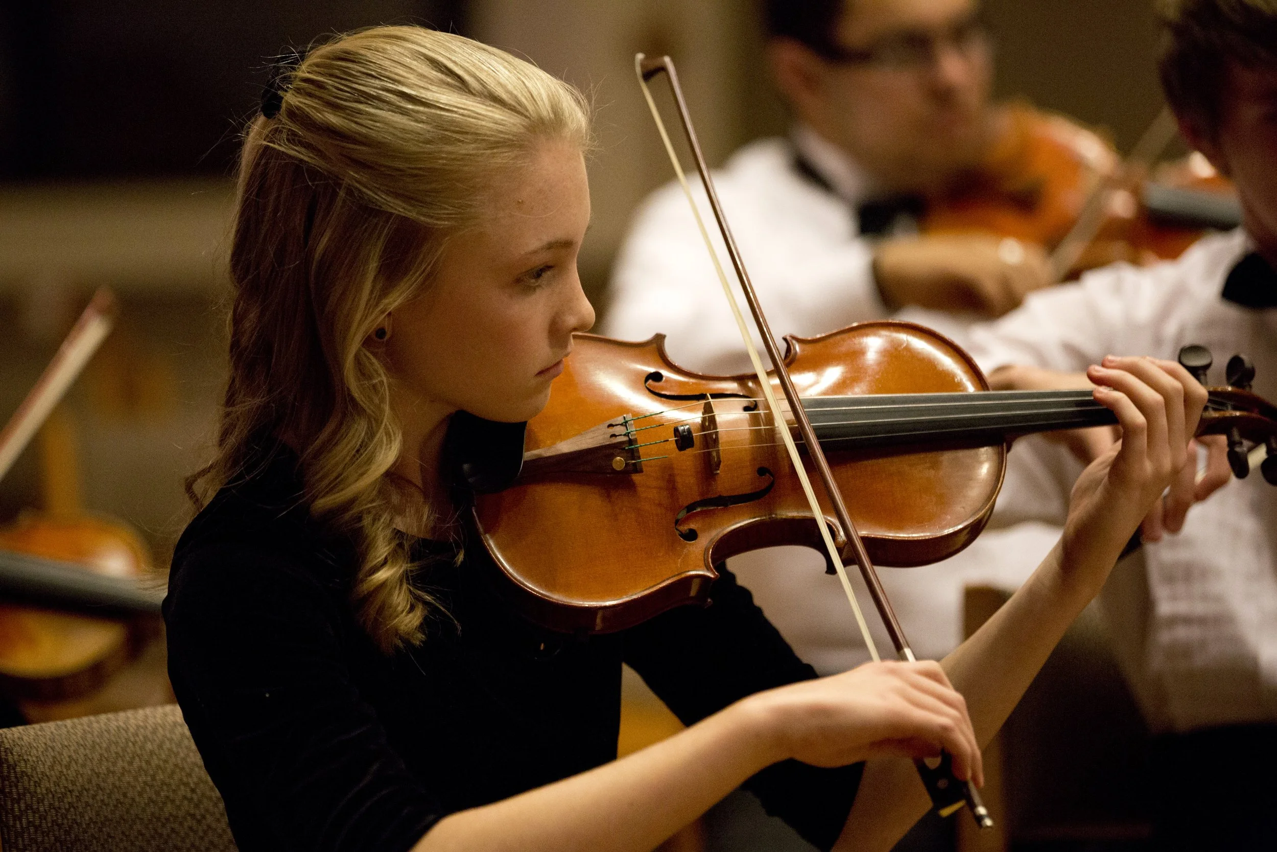 A young woman with blonde hair in a ponytail playing the violin during a performance.