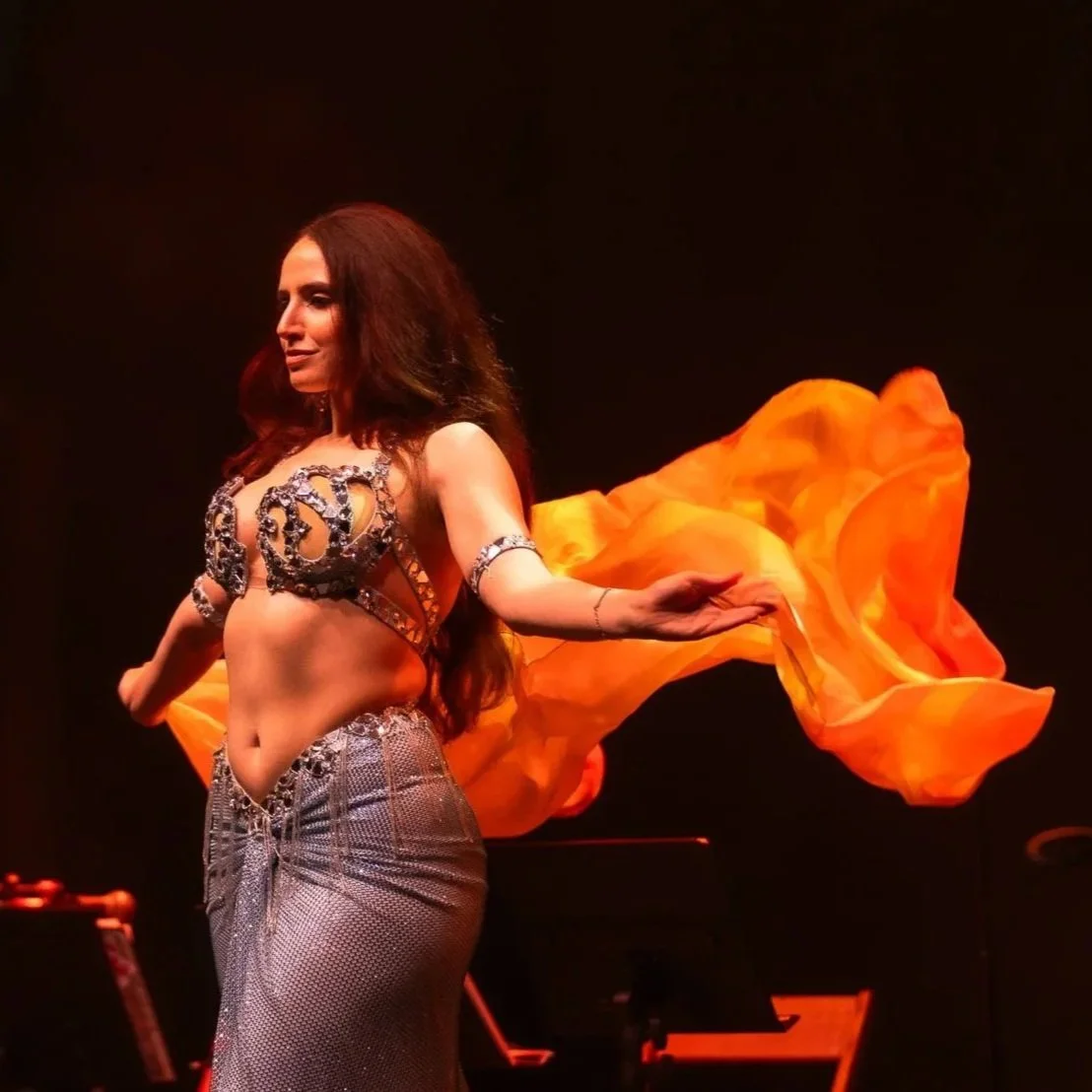A woman performing on stage in a belly dance costume, with orange fabric flowing behind her, in front of a dark background.