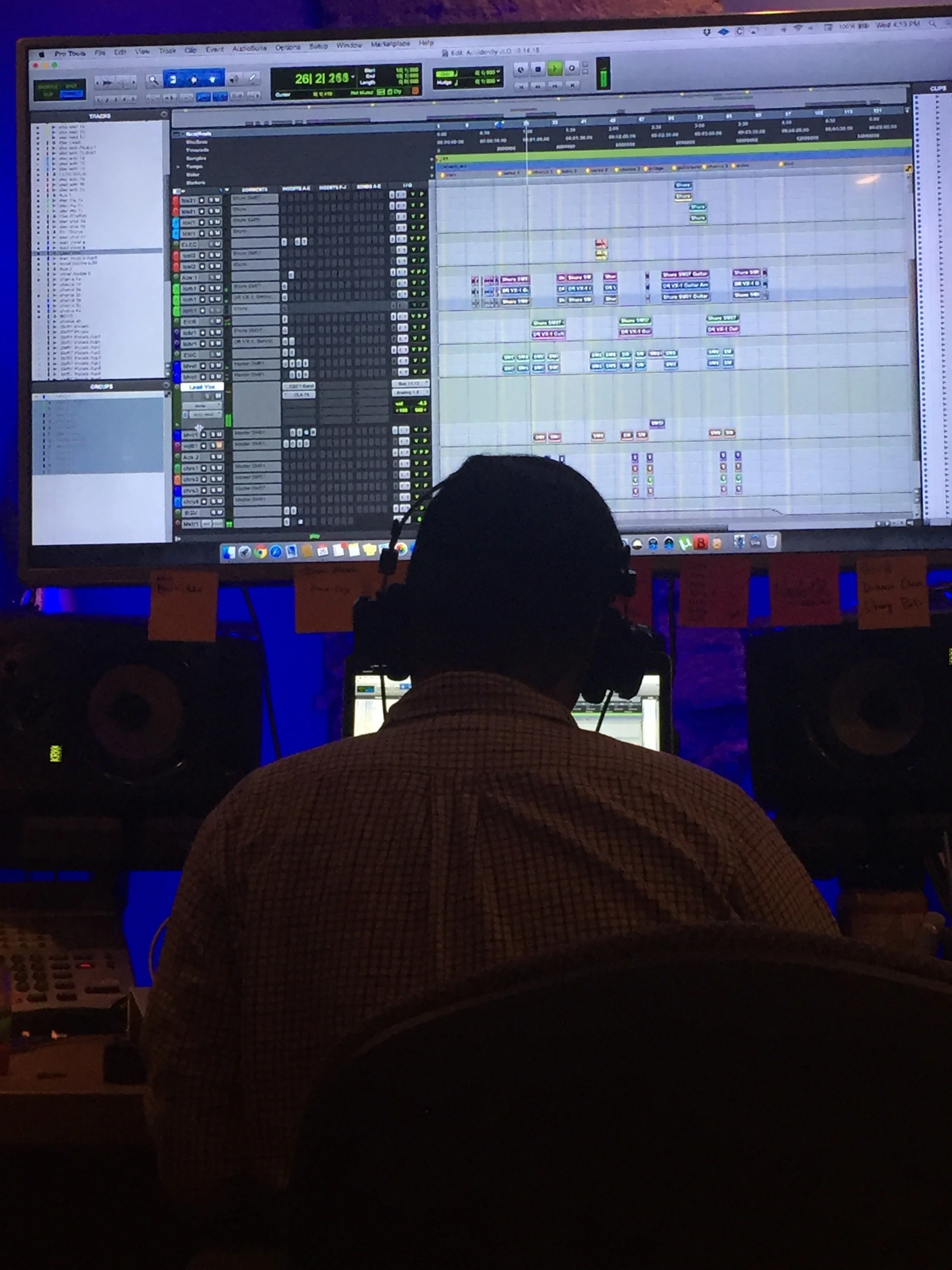 A person with headphones in a checkered shirt working on music production at a computer with a large screen showing digital audio workstation software in a dark room.