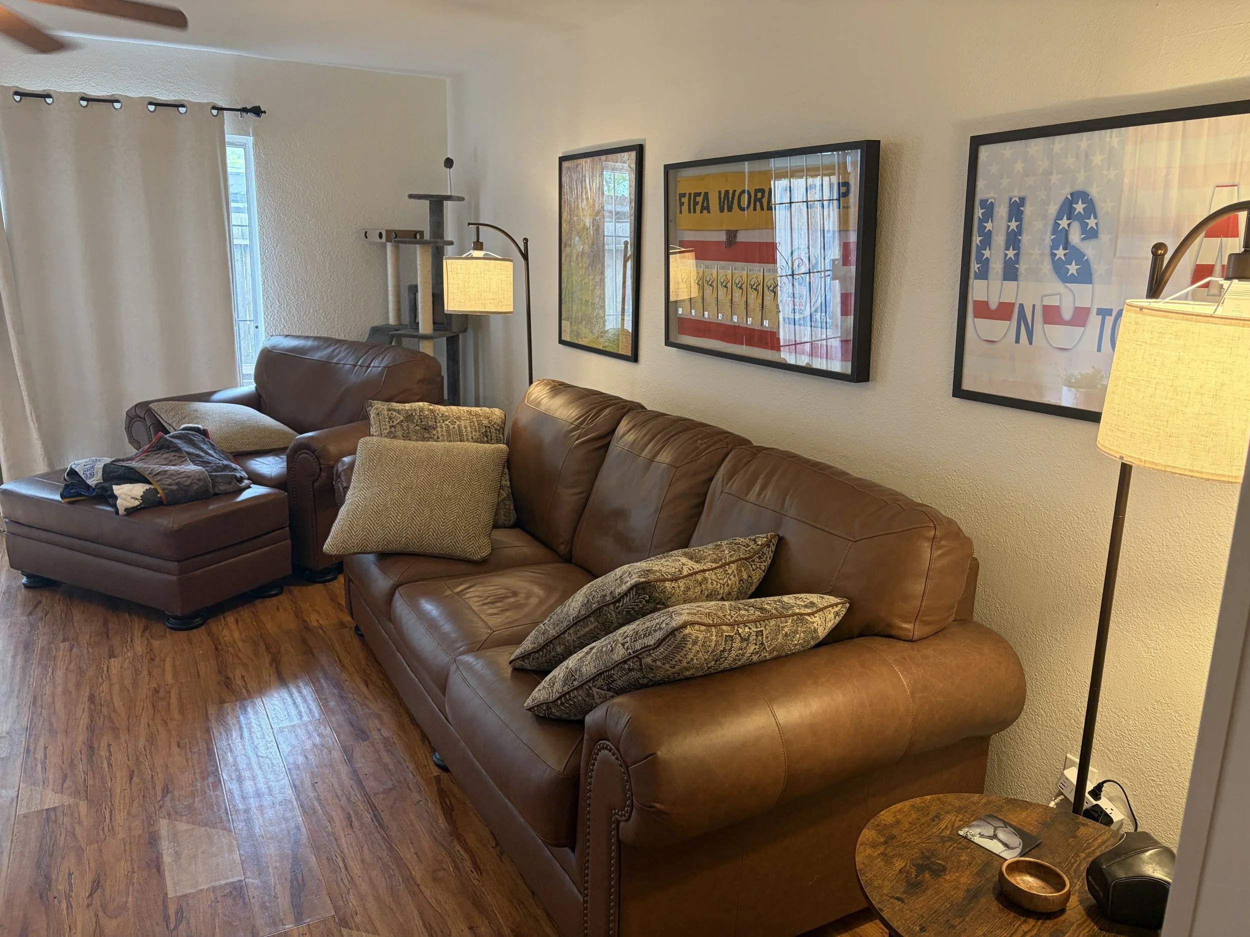 Living room with a brown leather couch, patterned throw pillows, a side table with a lamp, and hanging framed sports posters on a beige wall. There is a cat tree near the window with curtains, and hardwood flooring.