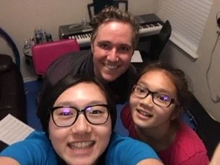 Three people smiling for a selfie in a music room with a keyboard and window blinds in the background.