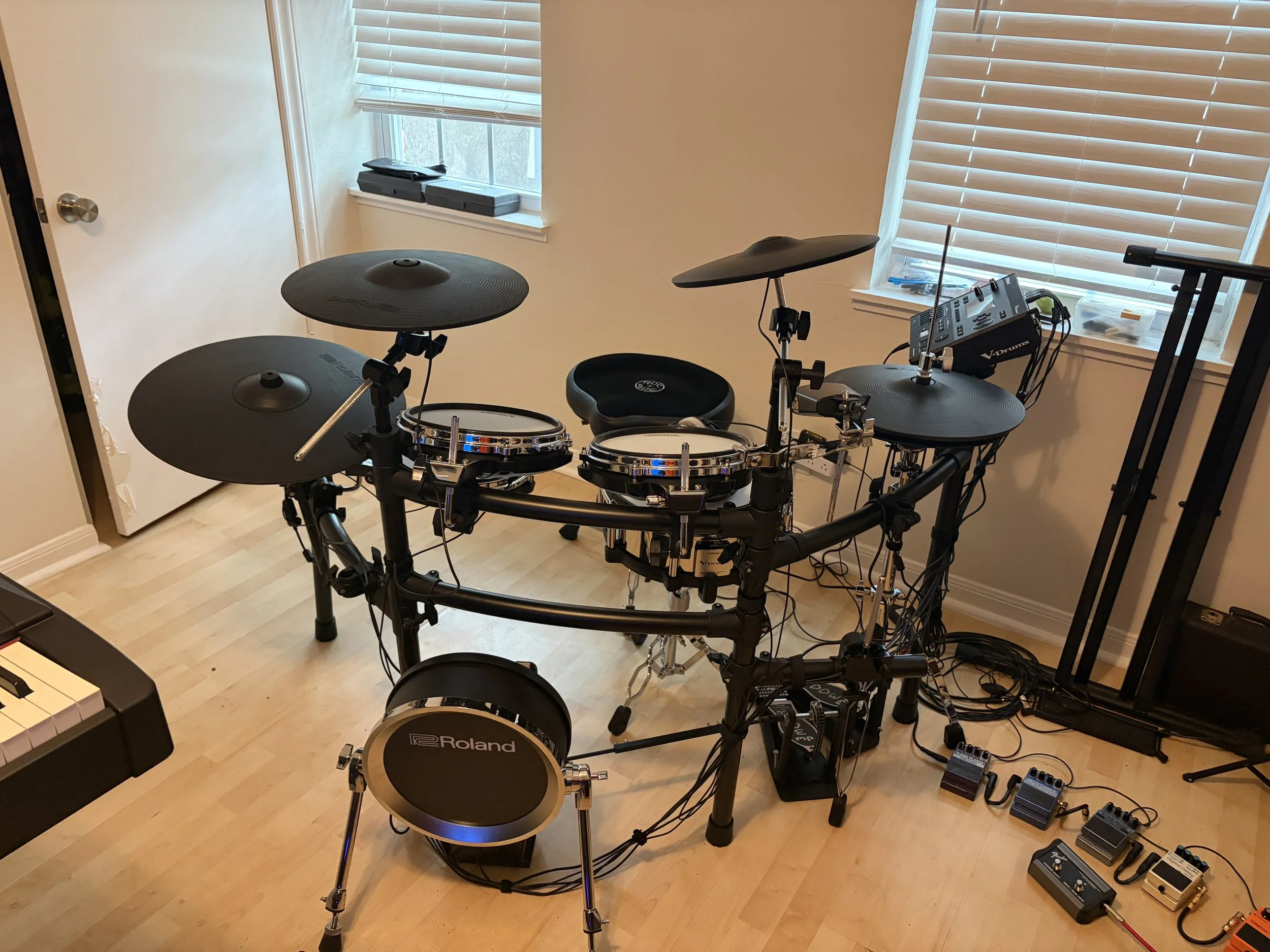An electronic drum set with cymbals, pads, and a rack, positioned near two windows with blinds, on a wooden floor in a room.