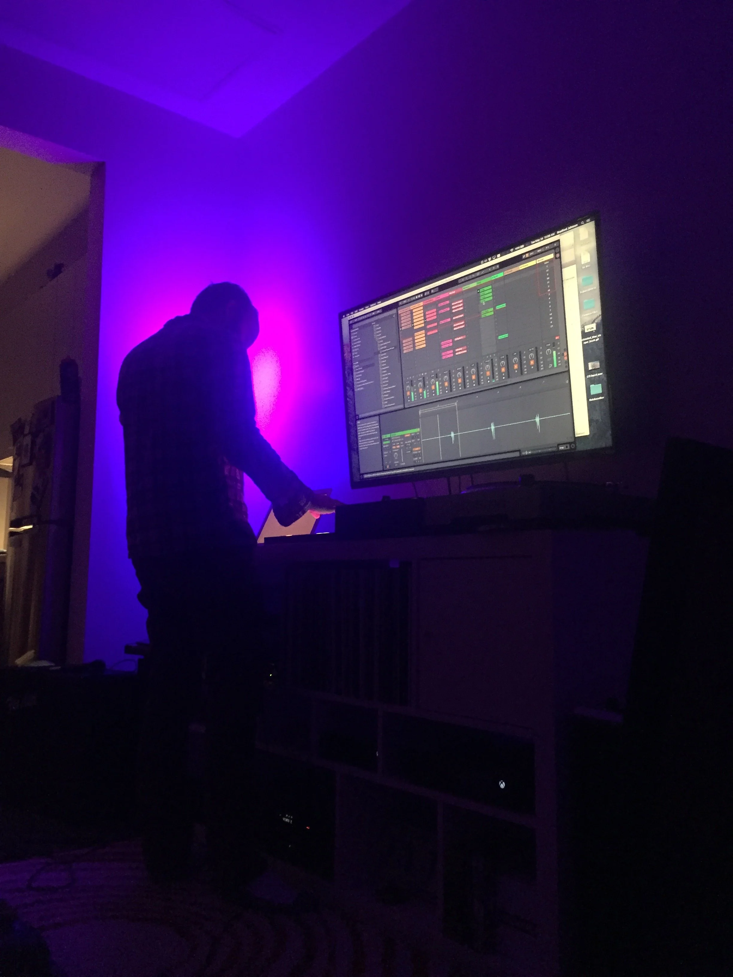 Person working at a music production setup with a large monitor displaying digital audio workstation software, purple ambient lighting illuminating the room