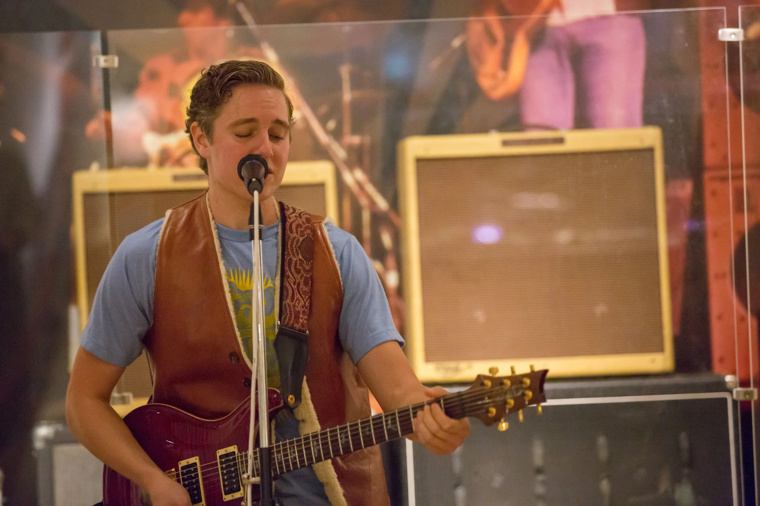 A young man with short hair singing into a microphone and playing an electric guitar in a music studio, with yellow amplifiers in the background.