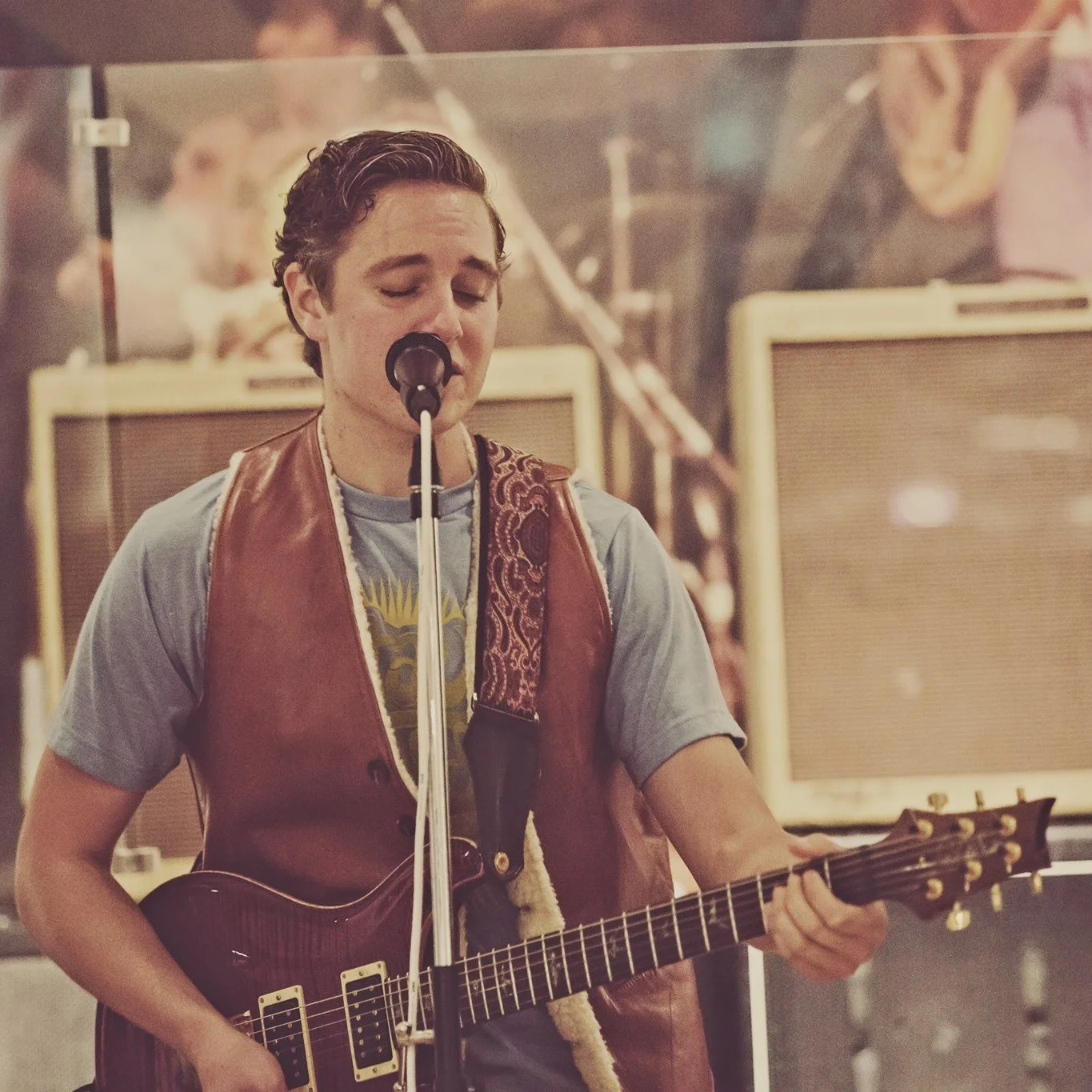A young man with closed eyes singing into a microphone while playing an electric guitar, in a music studio with amplifiers in the background.
