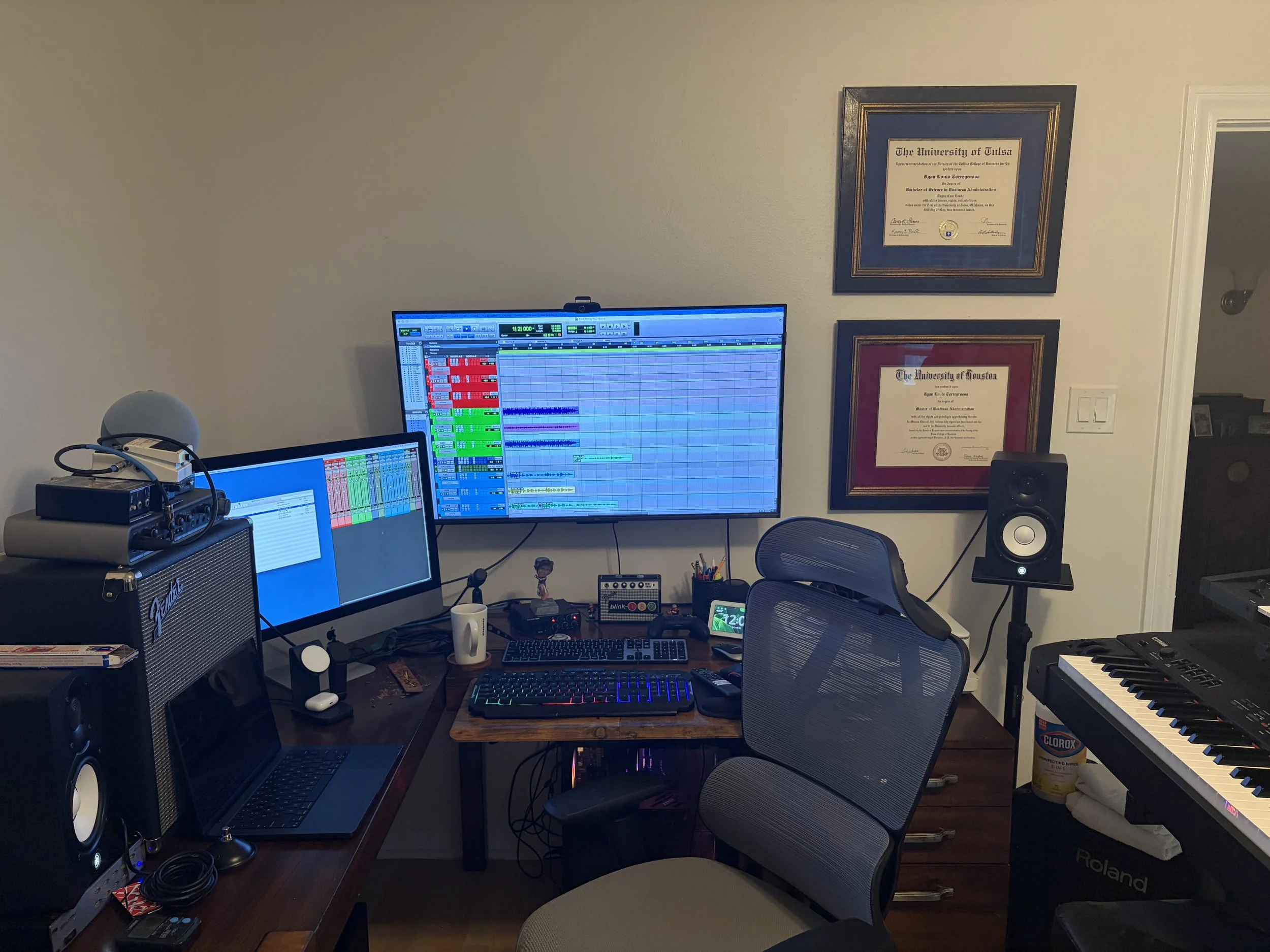 A music production workspace with multiple computer monitors displaying digital audio workstations and tracks, a keyboard, audio speakers, musical keyboards, audio equipment, and framed university diplomas on the wall.