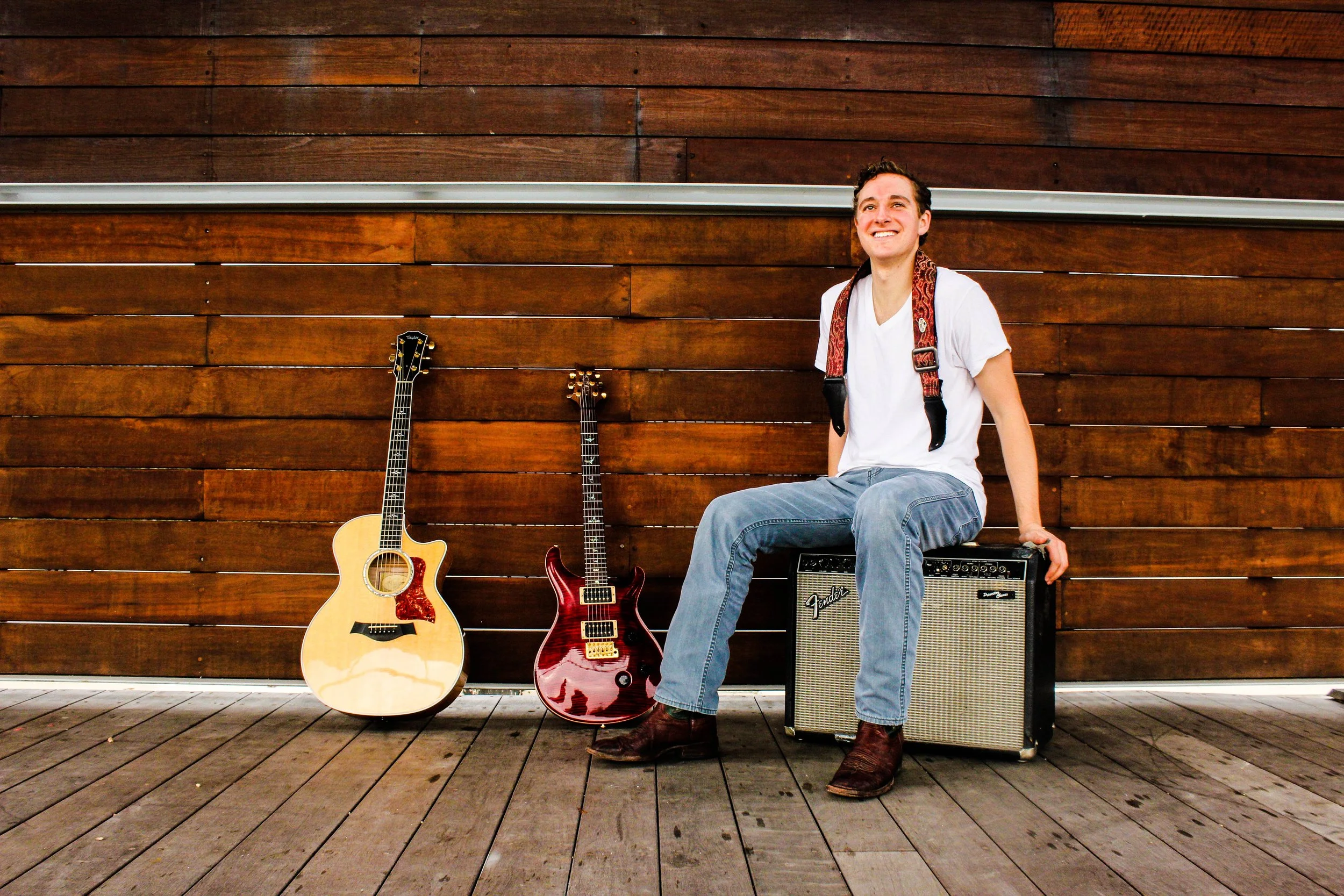A smiling man wearing a white t-shirt, jeans, and boots, sitting on an amplifier in front of a wooden wall, with two guitars leaned against the wall beside him.