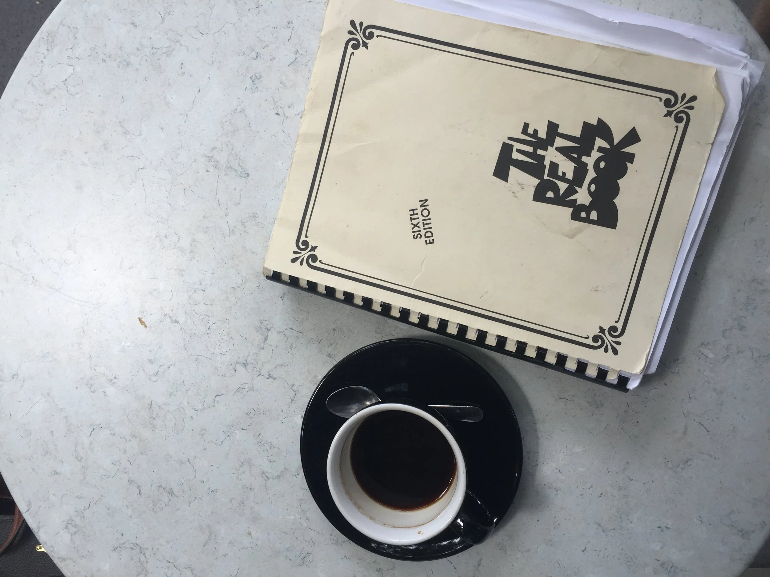 A white mug filled with black coffee and a smaller black cup of coffee placed on a round white and black saucer are on a marble table. Above them, there is a spiral-bound notebook with a cream cover labeled 'The Book' and 'Sixth Edition'