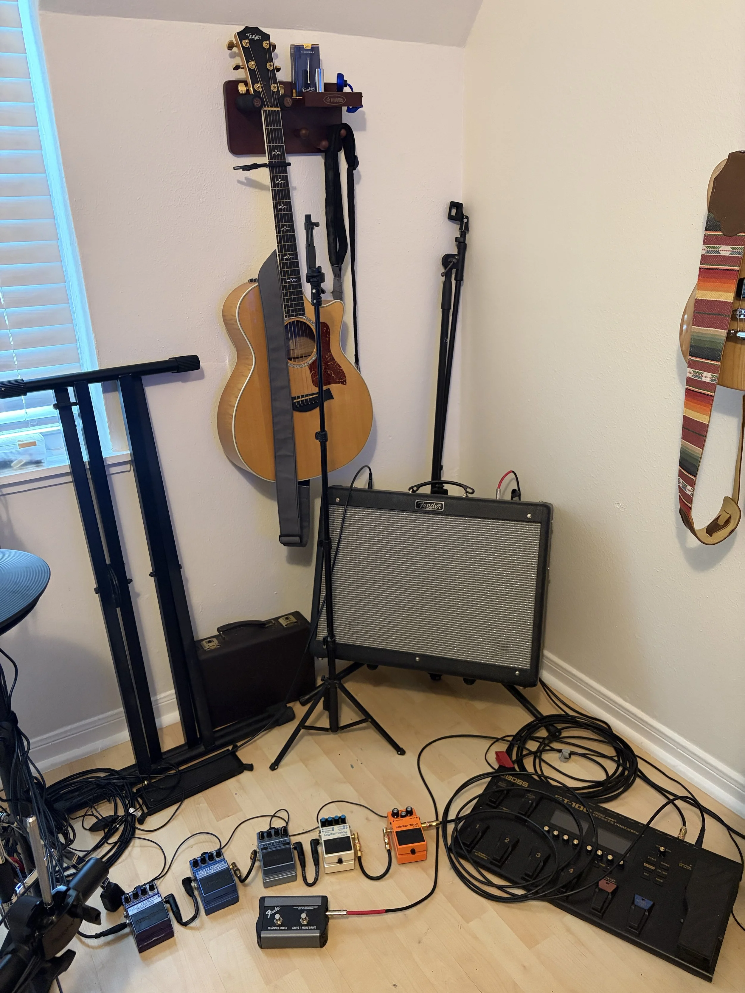 A corner of a room with musical equipment, including an acoustic guitar hanging on the wall, guitar effect pedals on the floor, a Fender amplifier, and various cables. There's also a music stand, a microphone, and part of a drum pad on the right.