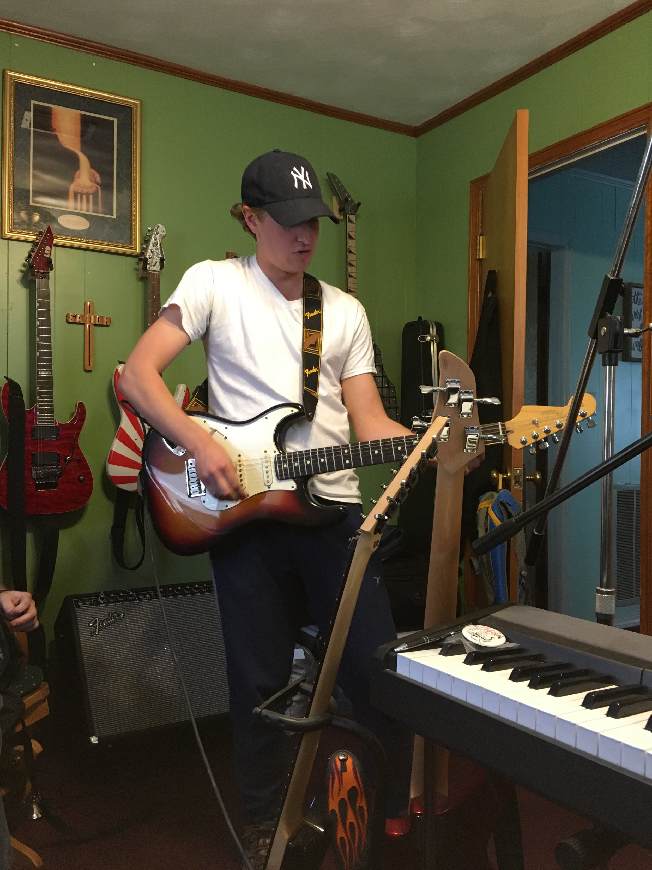 A young man wearing a black New York Yankees cap, white T-shirt, playing an electric guitar in a green-walled room with musical instruments and equipment. A framed picture featuring a fork and some electric guitars on the wall, and a fish-shaped cross hang behind him. There is a keyboard and a speaker on the floor.