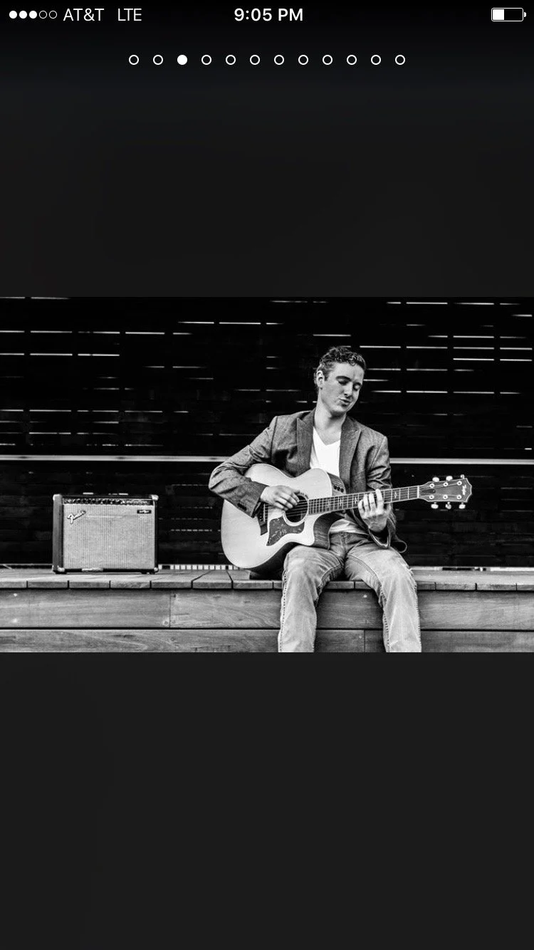 A young man sitting on a wooden platform playing an acoustic guitar, with an amplifier beside him and a dark patterned wall behind him, in black and white.