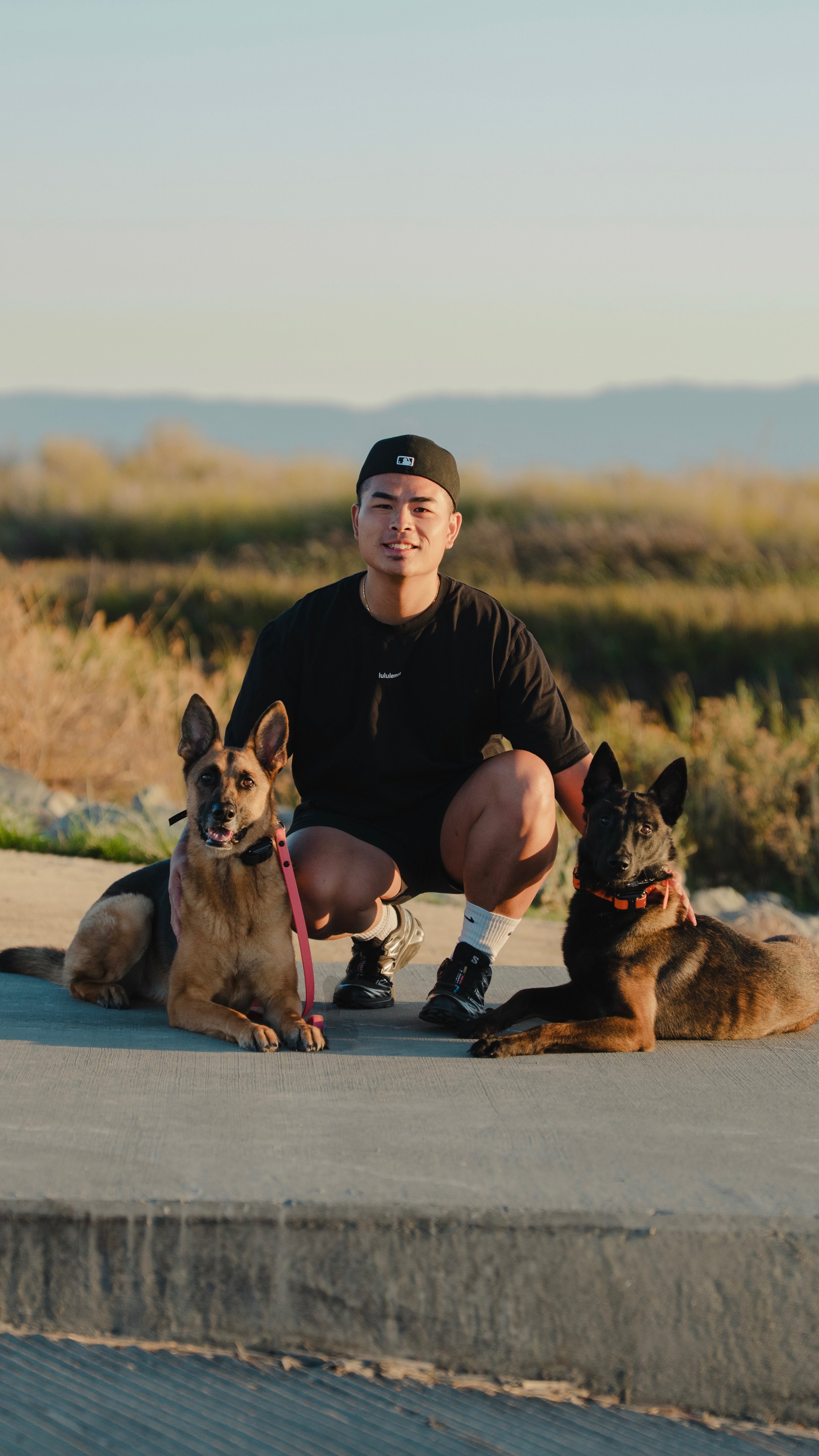 A young man crouching on a trail with two black and brown dogs, outdoors in the late afternoon with mountains in the background.