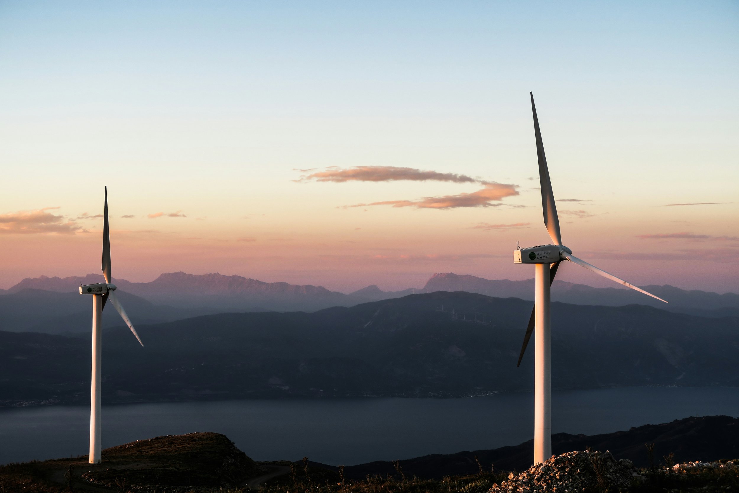 Two wind turbines on a mountain landscape at sunset with mountains in the background and a pinkish-orange sky.