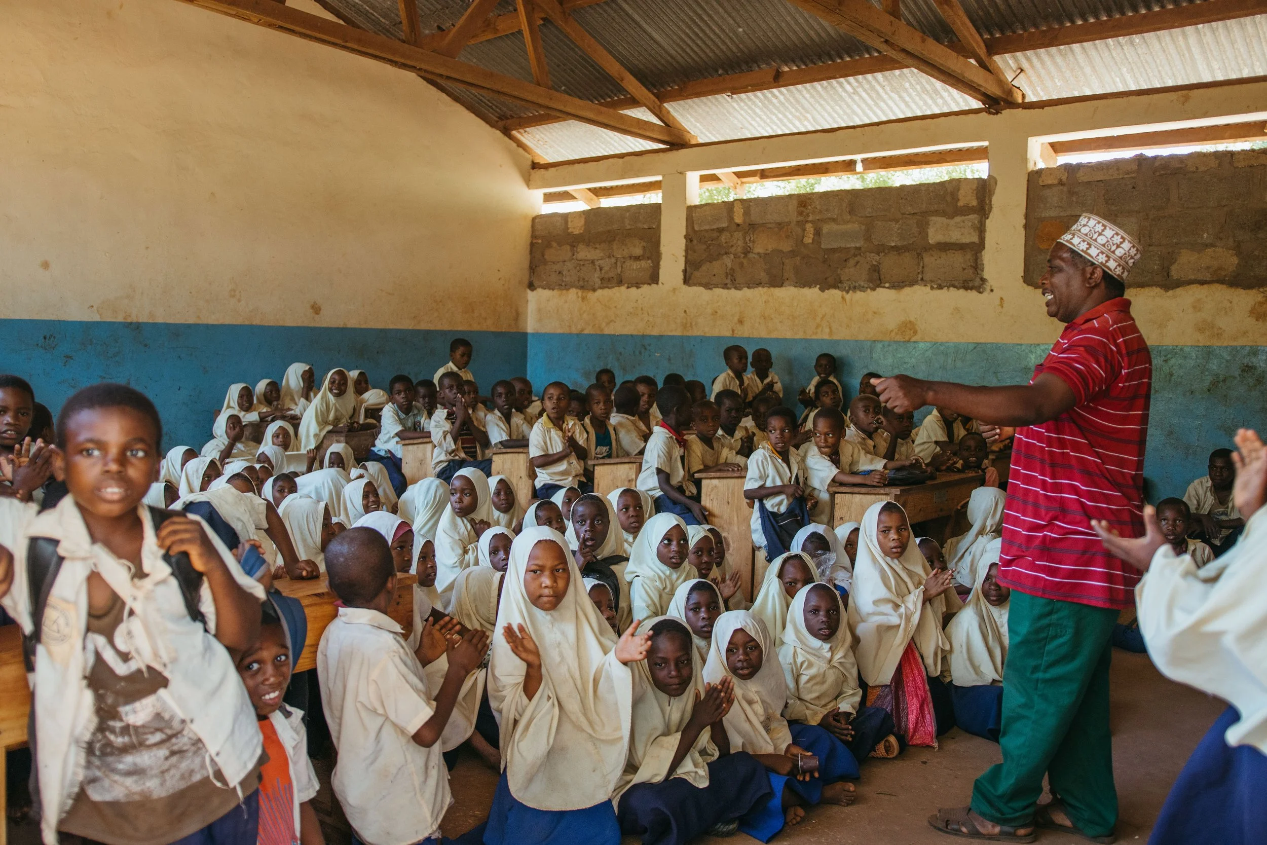 A classroom filled with young students in uniform, seated on wooden benches, with a teacher standing at the front, inside a simple building with a corrugated metal roof and a partially unfinished brick wall.