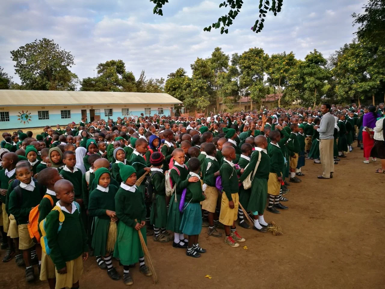 Large group of school children in green uniforms gathered outdoors, with adults supervising, on a school grounds with trees and a building in the background.