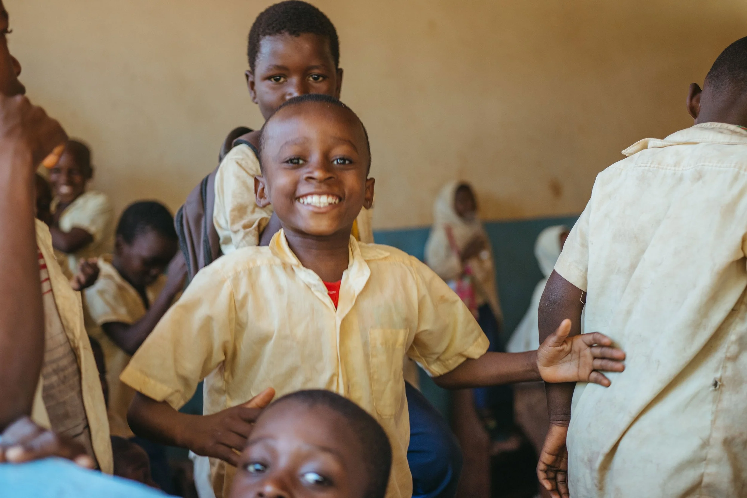 Smiling young boys in yellow school uniforms in a classroom, with other children in the background.