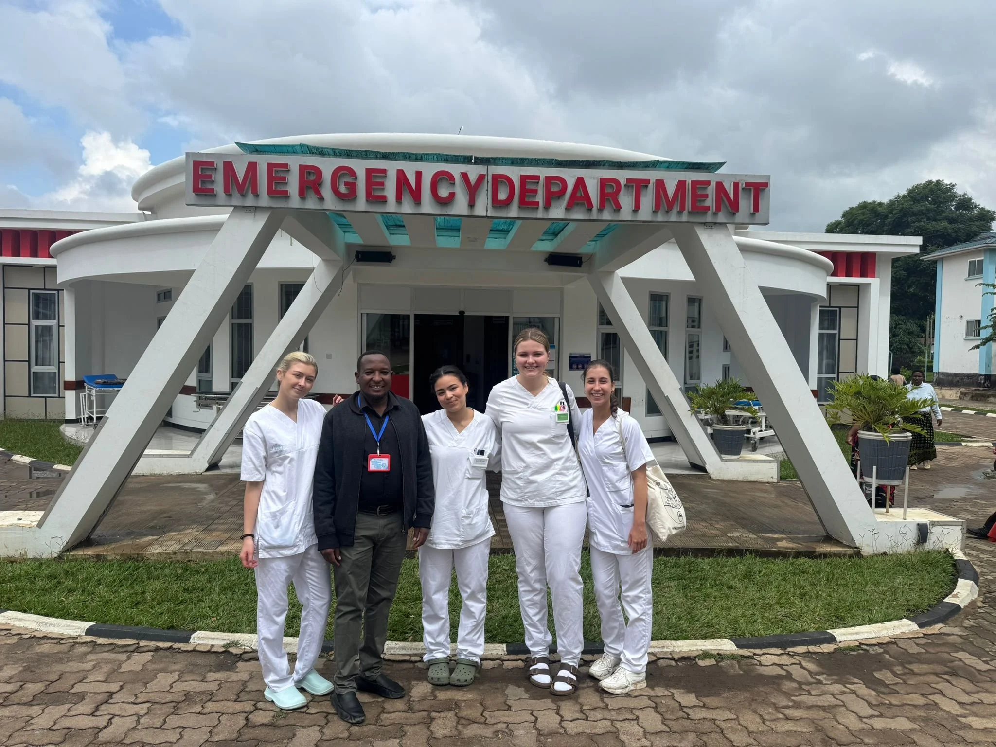 Group of five people standing in front of a building with a sign that reads 'Emergency Department'. They are wearing healthcare uniforms and casual clothing, smiling at the camera.
