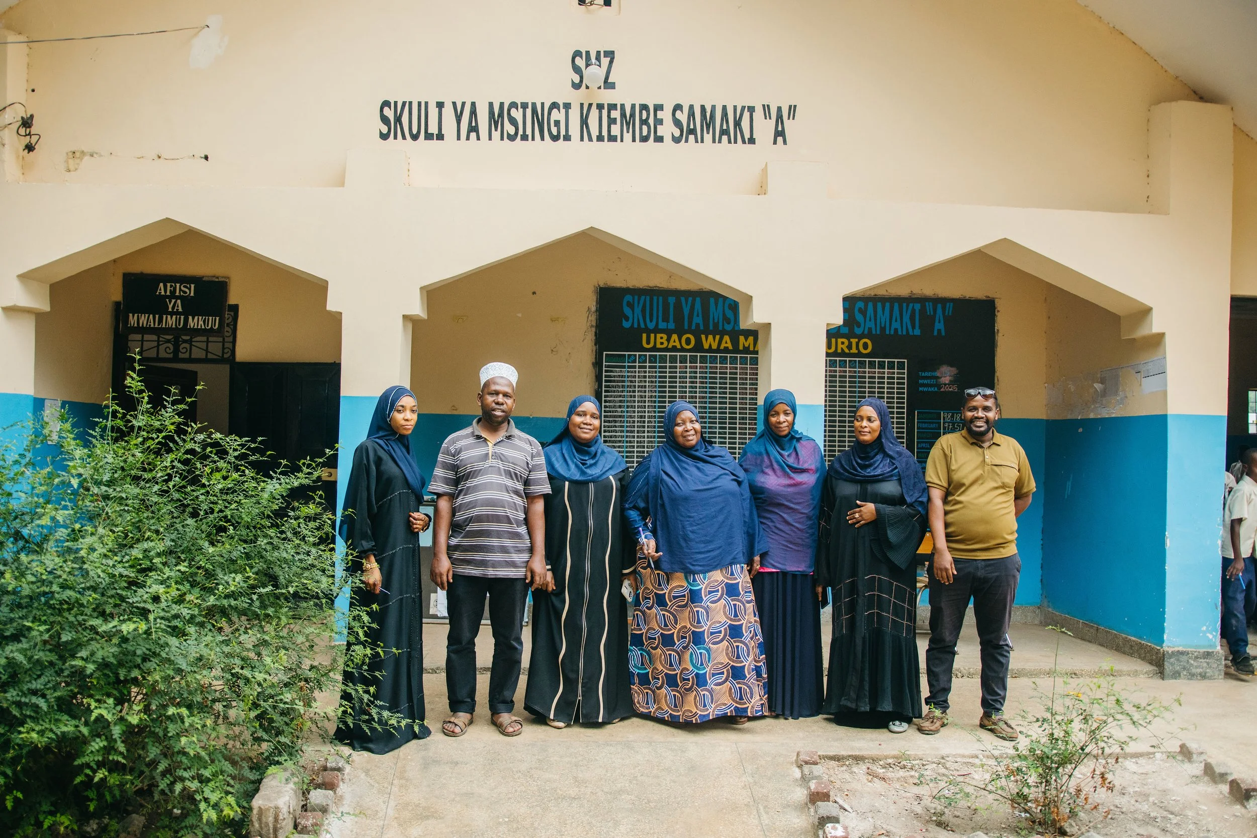 Group of seven people, four women and three men, standing outside a beige and blue building with signage, posing for a photo. The women are wearing headscarves, and the men are dressed in casual shirts. There are green bushes in the foreground.