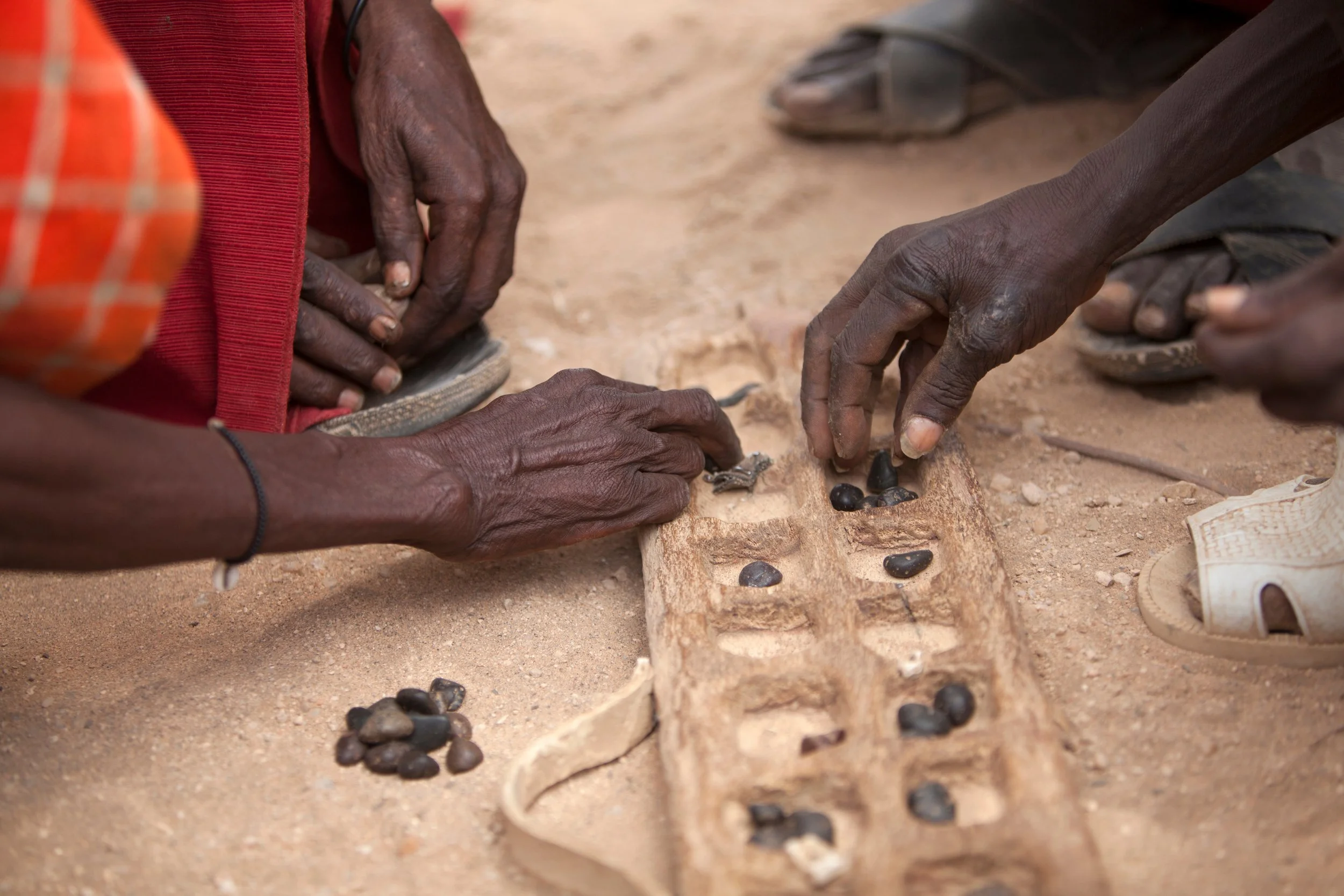 Close-up of four hands, three dark-skinned and one light-skinned, scooping black beans from a clay or stone container on a dirt ground. The scene suggests traditional food gathering or preparation.