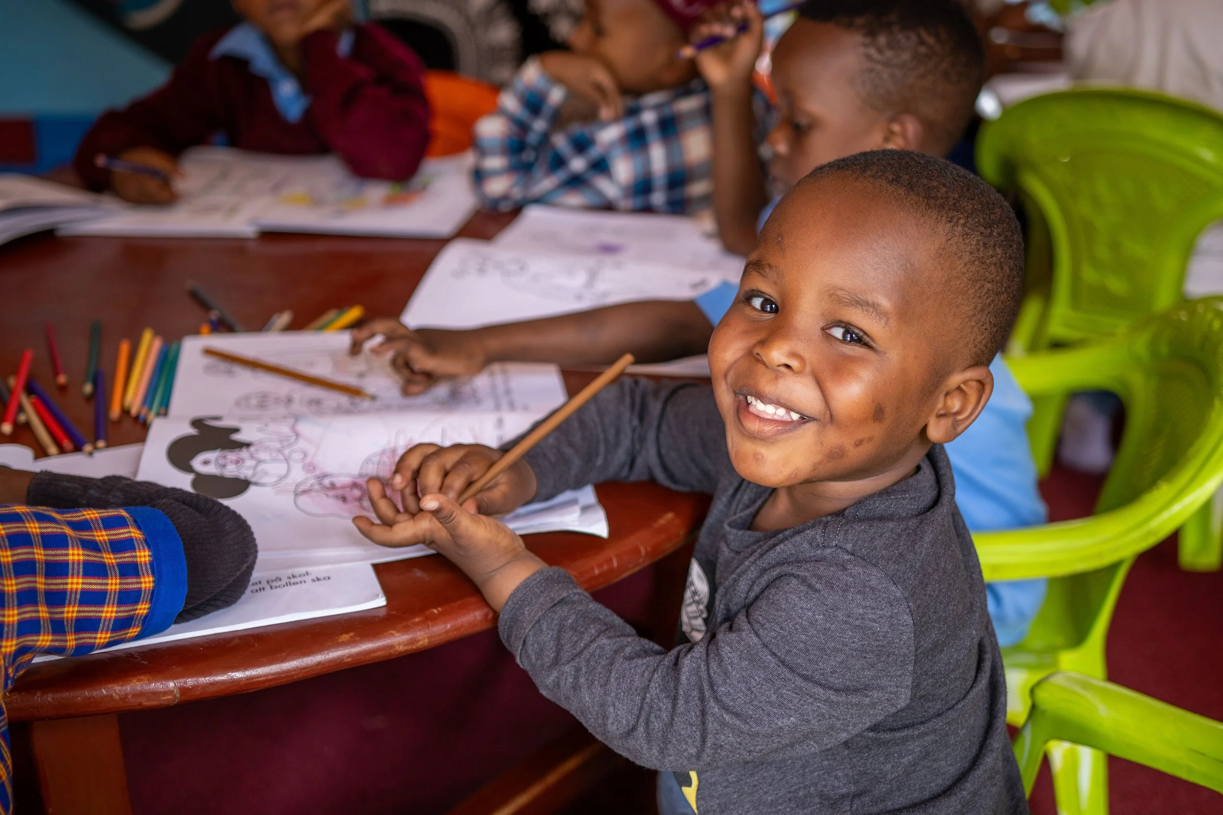 A young boy with a smile, sitting at a table with coloring pages, colored pencils, and crayons, engaged in an activity in a classroom or similar setting.