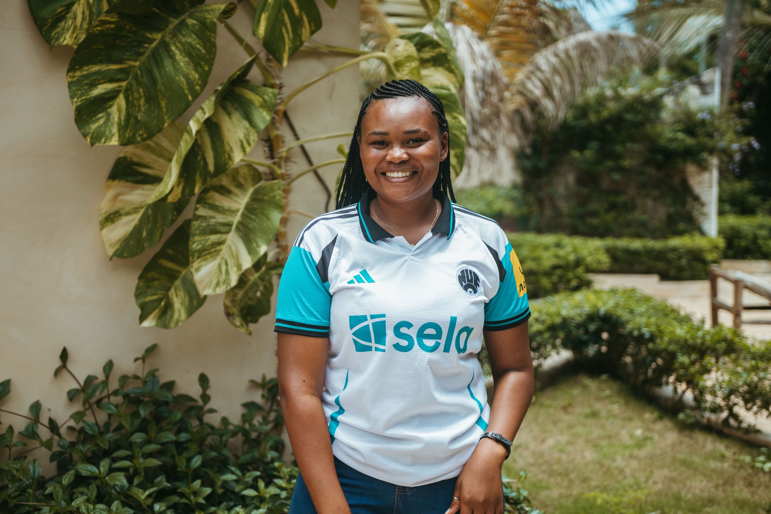 A woman stands outdoors smiling, wearing a sports jersey with the logo 'Sela' on it. She has braided hair and is surrounded by green plants and trees.