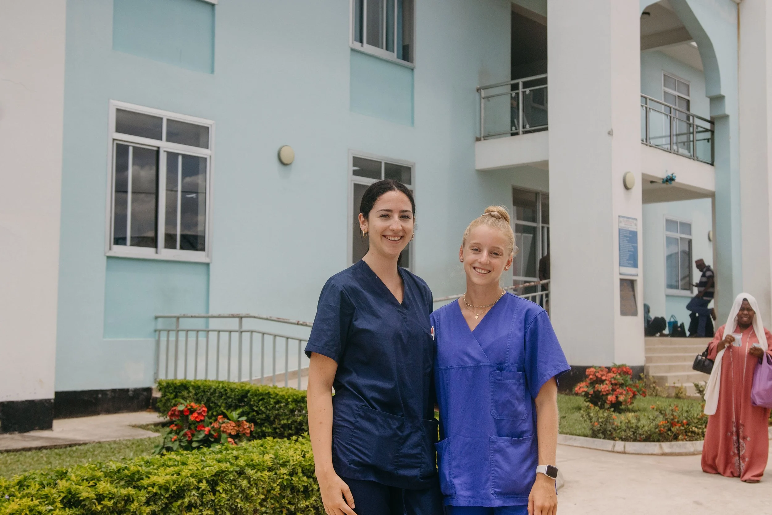 Two women, one with dark hair and the other with blonde hair, wearing blue medical scrubs, standing in front of a light blue building with multiple windows and balconies, smiling at the camera.