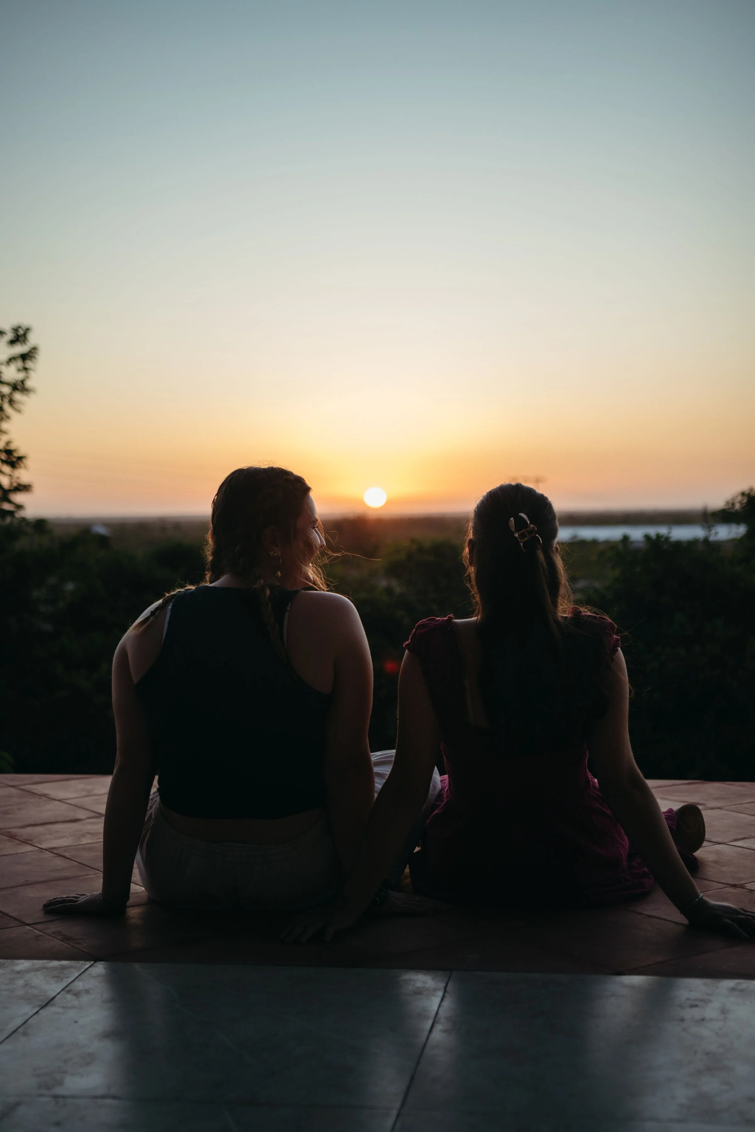 Two women sitting on a tiled surface outdoors at sunset, facing away from the camera, with a view of the sunset over a landscape with water and trees in the distance.
