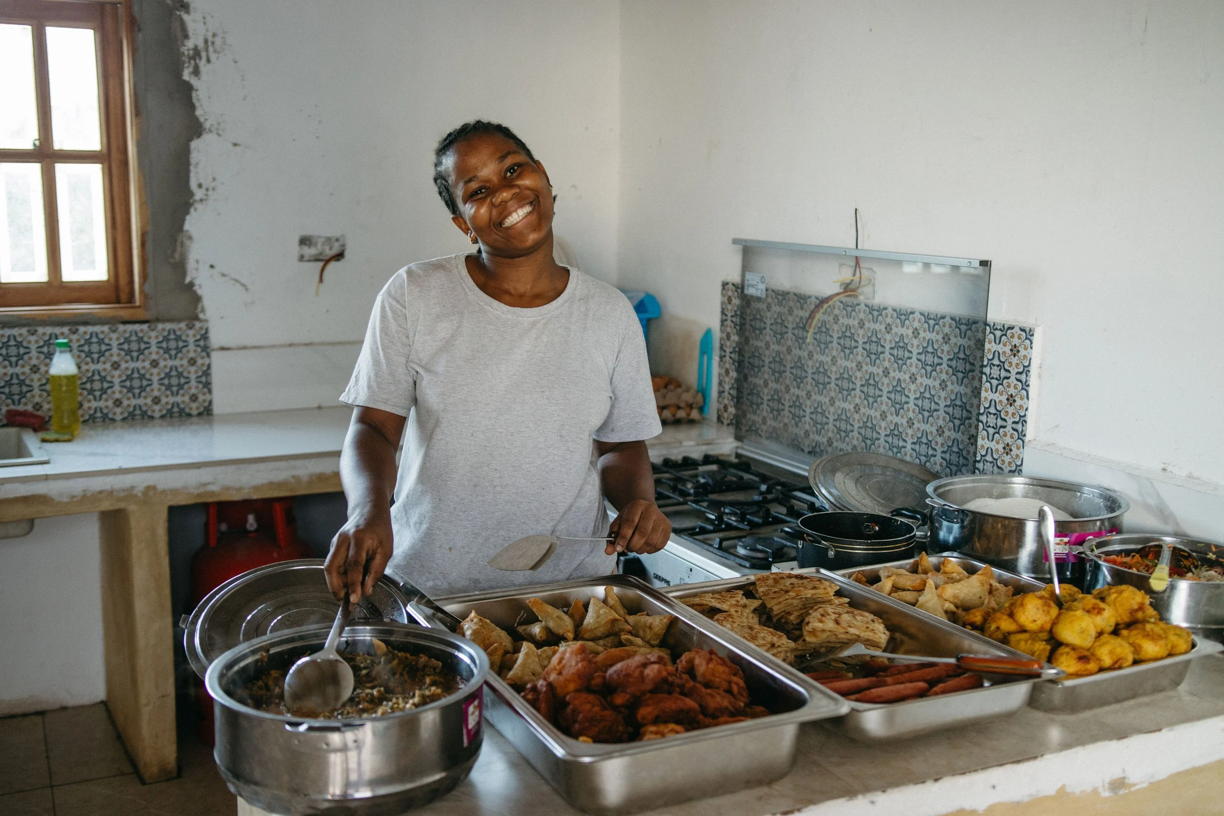 A woman smiling and serving food at a buffet in a kitchen with a window and patterned tile backsplash.