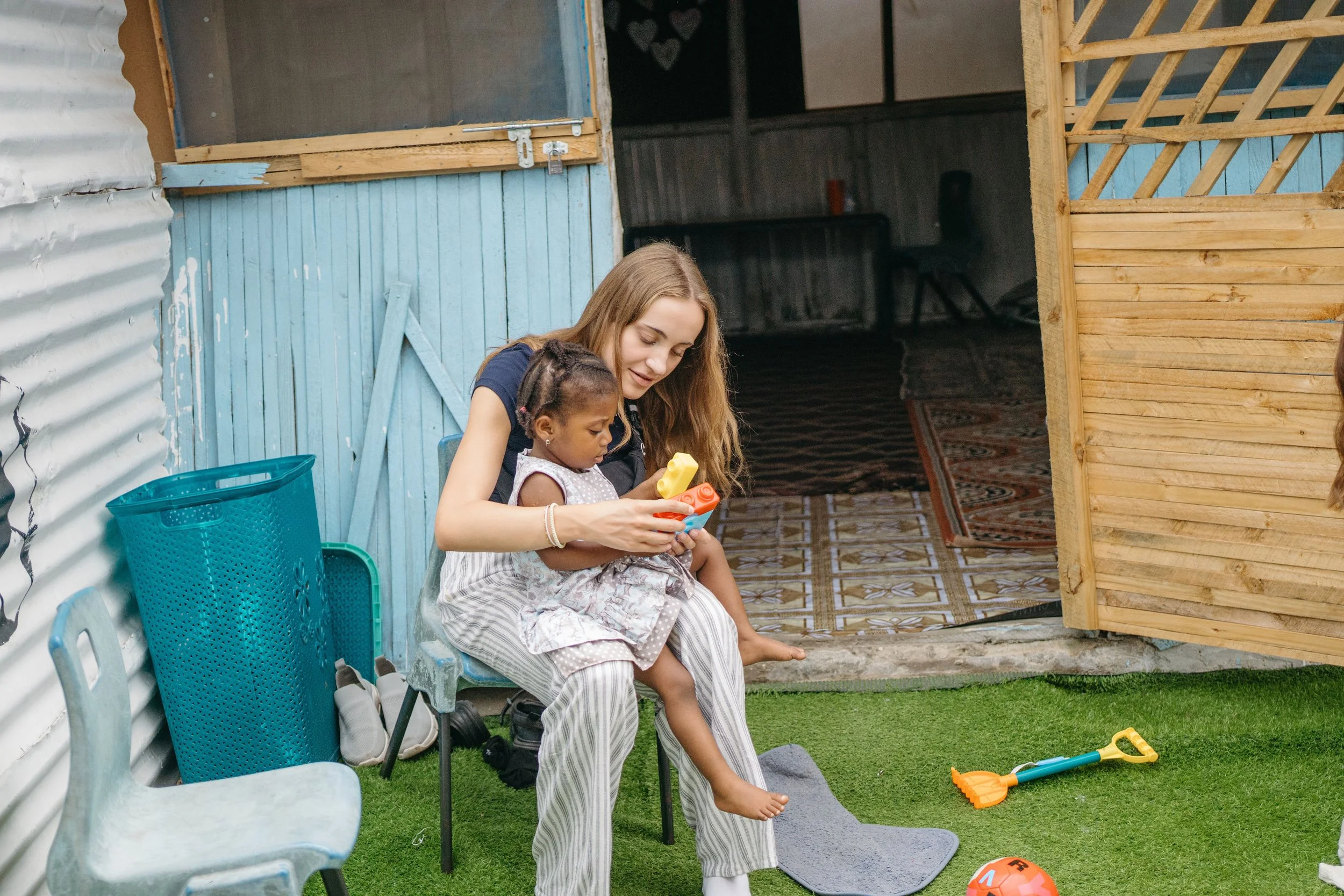 A woman and a young girl sitting on a chair outdoors, playing with toy blocks. The woman is smiling and the girl is focused on the blocks. There are various outdoor items around them, including a bright orange shovel, a soccer ball, and a green grass lawn.