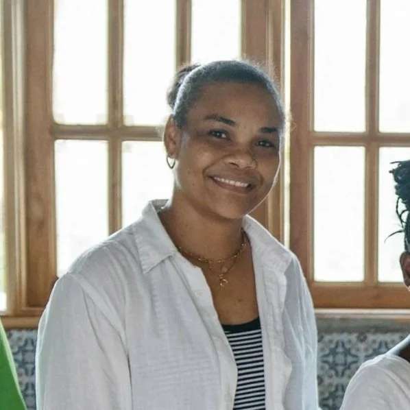 Smiling woman with dark hair tied back, wearing a white button-up shirt over a striped top, standing in front of wooden window panes.